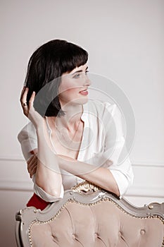 Studio portrait of attractive young woman with dark hair in white shirt and red pants leans on a classic chair on plain