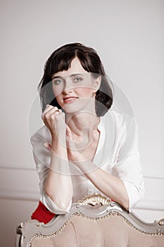 Studio portrait of attractive young woman with dark hair in white shirt and red pants leans on a classic chair on plain