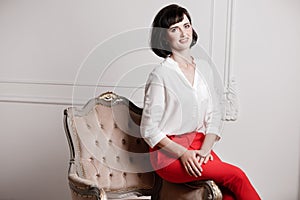 Studio portrait of attractive young woman with dark hair in white shirt and red pants sitting on classic chair on plain