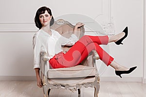 Studio portrait of attractive young woman with dark hair in white shirt and red pants sitting on classic chair on plain