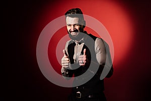 Studio portrait of a bearded man in a vest on a red background. The man`s hand shows a gesture of class