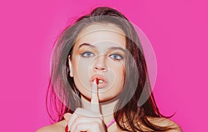 Studio portrait of beautiful elegant woman. Young lady showing shhh taboo sign with finger to lips over pink background