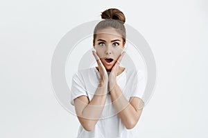 Studio portrait of beautiful european girl with bun hairstyle, holding hands on cheeks and expressing surprise or