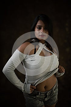 Studio portrait of a beautiful and young Indian Bengali female model in white cold shoulder top and jeans hot pant.