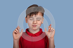 Studio portrait of a of a boy with closed eyes suggesting a desire and showing hands with crossed fingers on blue background in