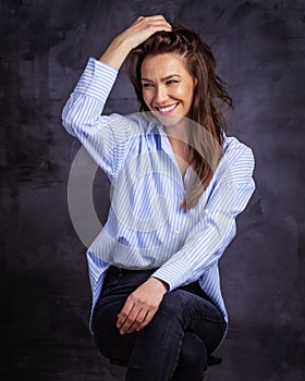 Studio portrait of a brunette haired attractive woman wearing blue shirt and black jeans while posing at isolated dark background