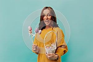 Studio portrait of charming brunette woman with curly hair wearing yellow pullover posing with basket with Easter eggs