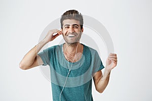 Studio portrait of cheerful hispanic guy in blue t shirt holding headphones with hand, listening music and dancing with