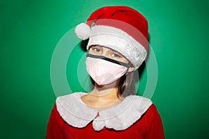 Studio portrait of child girl in santa costume wearing red hat and medical face mask on green background. Christmas concept.