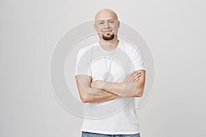 Studio portrait of confident good-looking bald bearded male standing with crossed hands over gray background, smiling