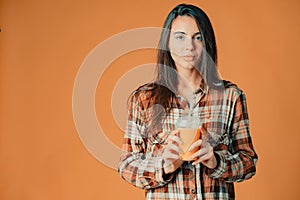 Cute caucasian young brunette girl holding plastic cup of orange juice on orange background