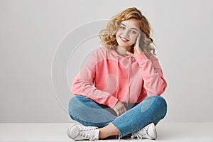 Studio portrait of cute female freelancer with curly hair sitting on floor with crossed lefs leaning head on hand and photo