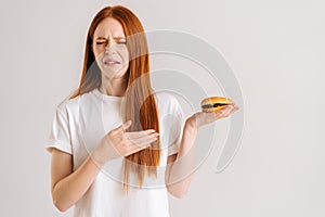 Studio portrait of dissatisfied pretty young woman with disgust pointing to burger, closed eyes, standing on white