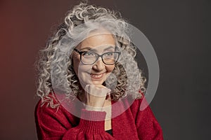 Studio portrait of an elderly woman 60-65 years old in a red sweater and glasses, gray curly long hair, on a colored background.
