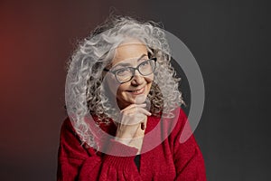 Studio portrait of an elderly woman 60-65 years old in a red sweater and glasses, gray curly long hair, on a colored background.