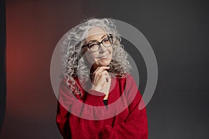 Studio portrait of an elderly woman 60-65 years old in a red sweater and glasses, gray curly long hair, on a colored background.