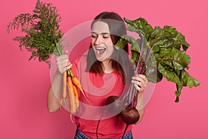 Studio portrait of emotional cheerful young vegetarian holding beet and carrots, opening mouth widely, looking at vegetables,