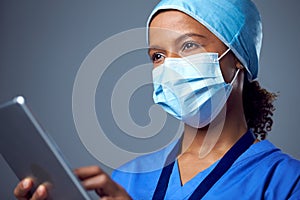 Studio Portrait Of Female Surgeon Wearing Scrubs And Face Mask Using Digital Tablet