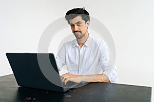 Studio portrait of friendly young man in shirt working on laptop computer, typing on keyboard, looking at camera sitting
