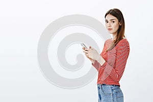 Studio portrait of gorgeous feminine european woman with brown hair, standing in profile, holding smartphone and looking