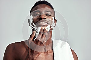 Studio portrait of handsome black guy with naked torso applying cream on his face. Afro American man with towel on his