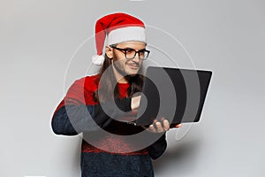 Studio portrait of handsome thoughtful smiling guy, holding laptop and typing on keyboard. Wearing Santa Claus hat. Christmas