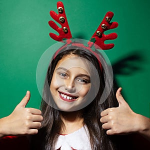 Studio portrait of happy child girl wearing reindeer horns and Santa costume showing thumbs up on green background.