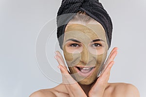 Studio portrait of happy young woman applying facial cosmetic green clay organic mask on her face, wears black towel on hair.