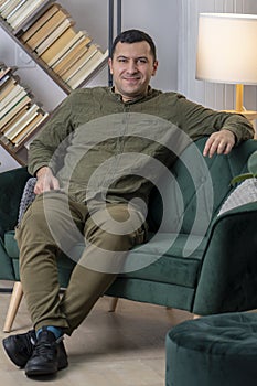 Studio Portrait of a man 30-35 years old, sitting on a sofa, shelves with books and a lamp.