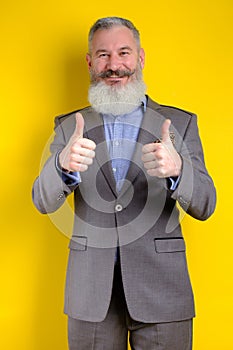 Studio portrait mature businessman dressed in gray suit shows thumbs up, successful business concept, yellow background