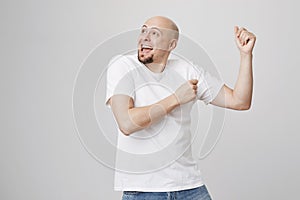 Studio portrait of positive overwhelmed bald caucasian man with beard dancing with raised hands, looking aside and