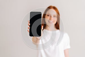 Studio portrait of positive redhead young woman with wide smile showing blank screen mobile phone and looking at camera,