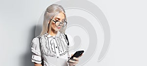 Studio portrait of pretty girl with blonde hair, using smartphone, wearing eyeglasses on white background, panoramic banner view