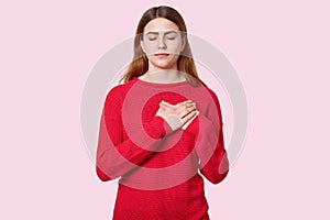 Studio portrait of restful impressed woman keeps both hands near heart, feels touched to hear heart piercing story, expresses good