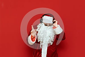 Studio portrait of Santa Claus in sunglasses listening to music in headphones and dancing on a red background. Santa party-goer in photo