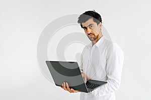 Studio portrait of serious confident young man holding in hand opened laptop computer and looking at camera standing on
