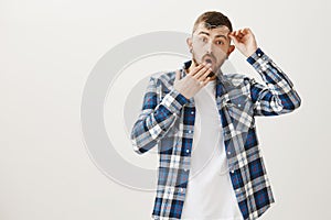 Studio portrait of shocked worried attractive man with beard, taking off glasses and holding eyewear near forehead