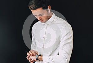 Studio portrait of smart young man wearing white shirt, looking at smartwatch posing over black background hurry up for meeting.