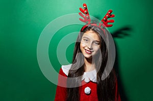 Studio portrait of smiling child girl wearing reindeer horns and Santa costume on green background with copy space.