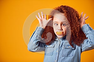 Studio Portrait Of Smiling Girl Pulling Funny Face With Orange For Mouth Against Yellow Background