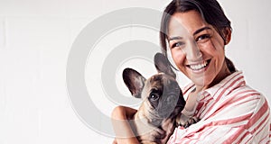 Studio Portrait Of Smiling Young Woman Holding Affectionate Pet French Bulldog Puppy