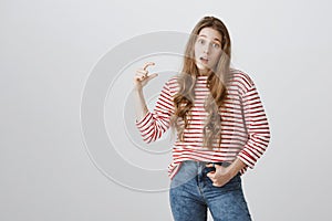 Studio portrait of surprised careless caucasian girl with blonde hair in striped sweater shaping something small over