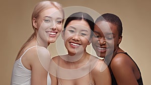 Studio portrait of three happy multiethnic ladies laughing to camera, enjoying friendship, hugging over beige background