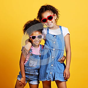 Studio portrait two mixed race girl sisters wearing funky sunglasses Isolated against a yellow background. Cute hispanic
