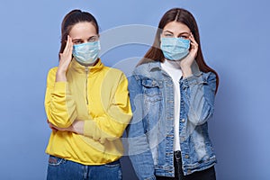 Studio portrait of two young girls with headache, holding hands on temples, wearing medical flu mask and posing iolated over blue