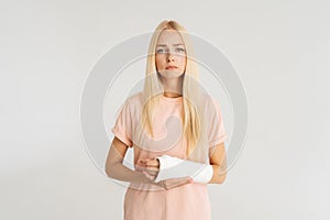 Studio portrait of unhappy injured blonde young woman with broken arm wrapped in plaster bandage looking at camera