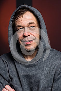 Studio portrait of a 40-50-year-old laughing man in a gray hoodie on a neutral background with a hood on his head, looking away.