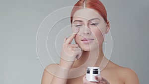 Studio portrait of young, beautiful and natural redhead woman applying skin care cream. Face lifting, cosmetics and make