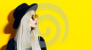 Studio portrait of young, beauty blonde girl wearing black hat and sunglasses. Looking away at empty background of yellow.