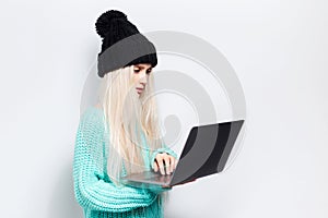 Studio portrait of young blonde girl using laptop on white background. Wearing black hat and cyan sweater.